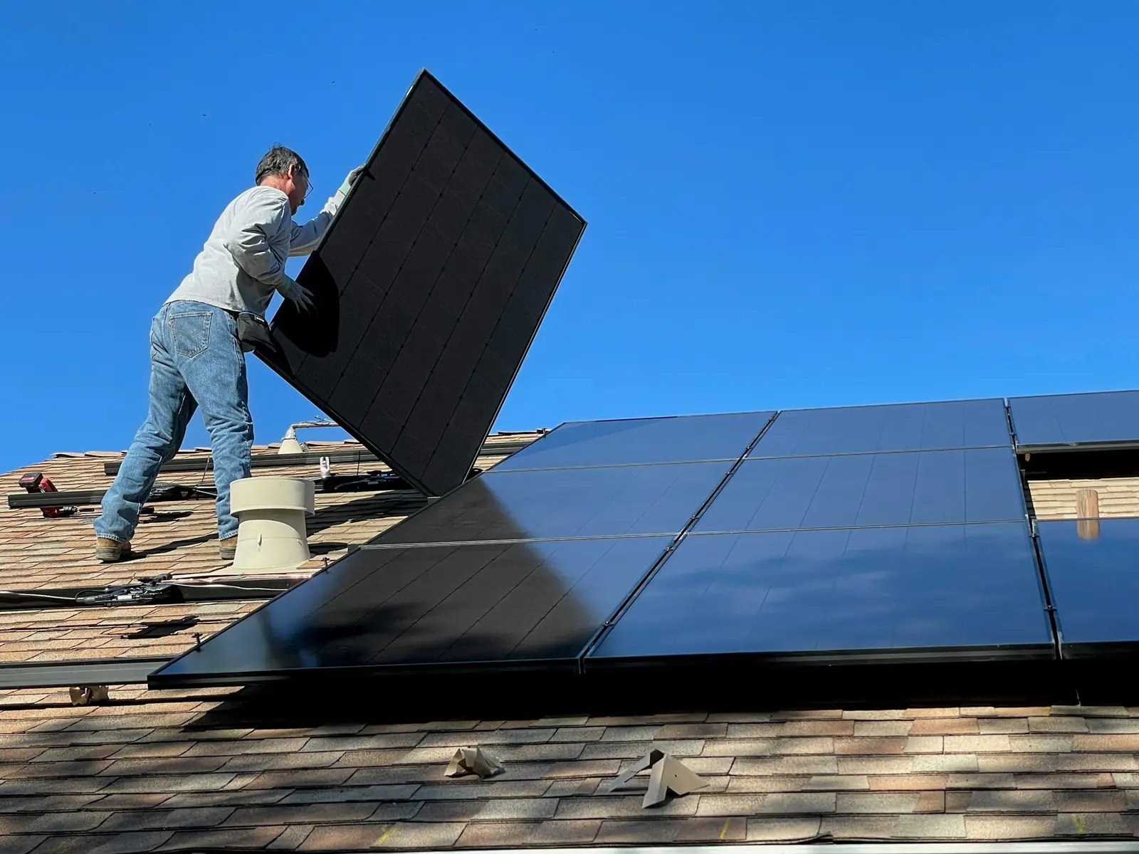 Technician carrying a solar panel across a shingle roof next to installed panels