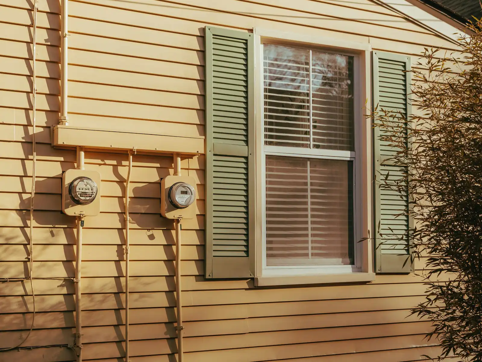 Two electric meters on the yellow siding of a house beside a window