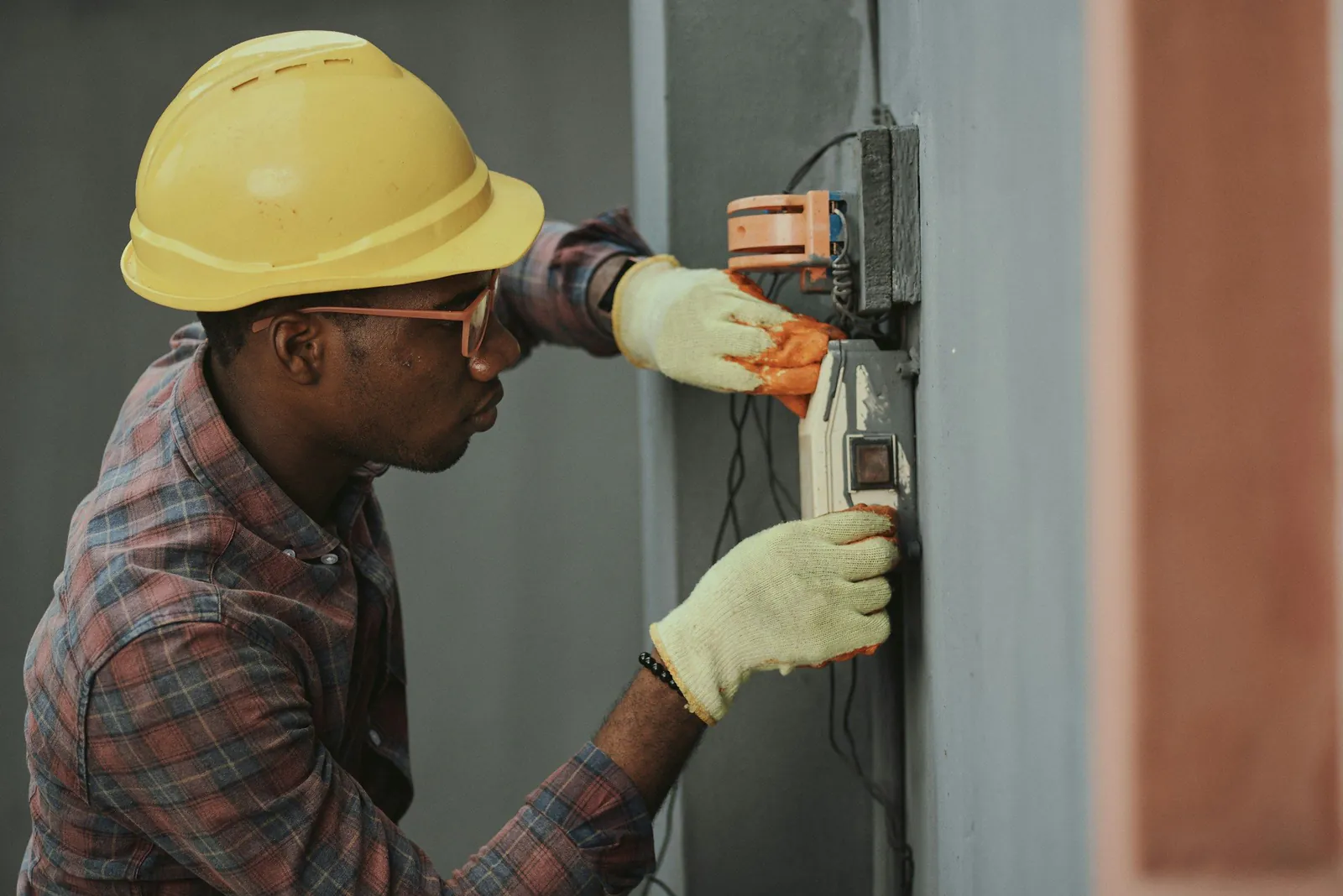 Electrician in a hard hat and gloves wiring a wall-mounted electrical box