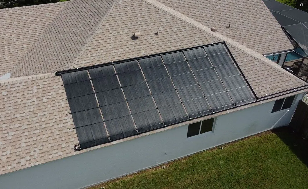 Aerial view of black pool heating collectors on a grey shingle roof beside a lawn